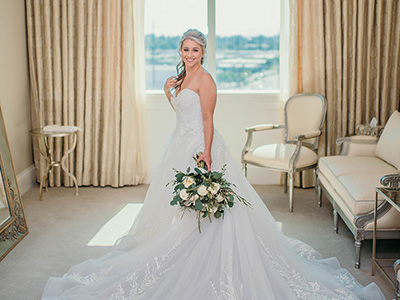 A person wearing a wedding gown stands alone in a bright dressing room holding a bouquet, framed by a large window, chandelier, and classic upholstered furniture.