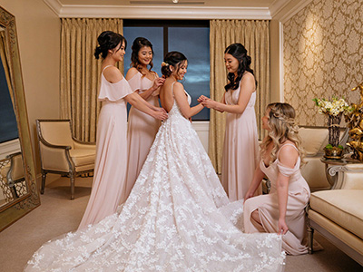 Several attendants gather around a person wearing a wedding gown in a dressing room, carefully adjusting the fitted bodice and long lace train beneath warm chandelier lighting.