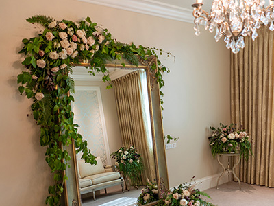 A large decorative mirror adorned with lush greenery and pale flowers stands in an elegant dressing room, with soft natural light streaming across the floor.