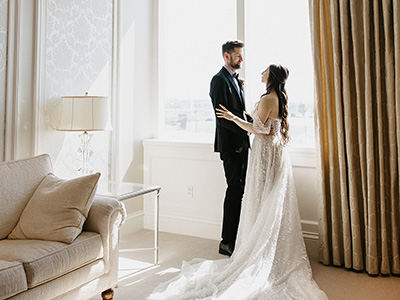 Two people stand facing each other near a large window in a refined suite, with sunlight illuminating a long flowing gown and neutral-toned furnishings.
