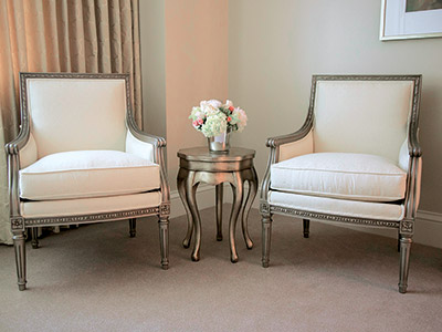 Two white chairs on either side of a silver end table holding flowers.