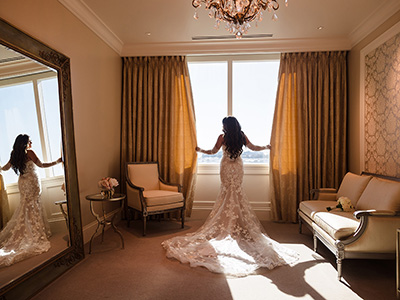 A person wearing a wedding gown stands with arms extended while facing a bright window in a refined suite, with the gown’s lace train flowing across the carpeted floor.