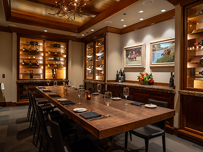 Private dining room with a long wooden table, wine shelves, framed artwork, and warm wood lighting