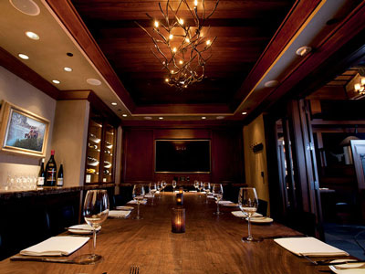 A room with a long dining table, chandelier, built-in wine cabinets, counter, and a tv monitor.