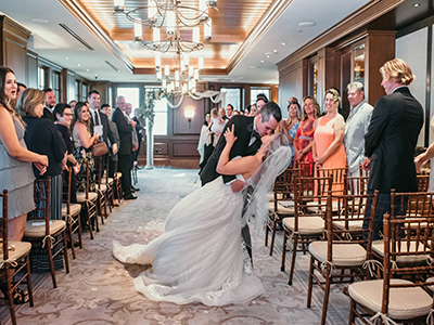A wedding ceremony moment inside a wood-paneled room where a couple shares a kiss in the aisle as guests stand on either side beneath warm chandelier lighting.