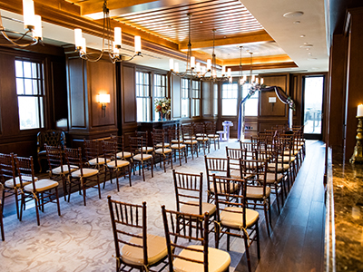An empty indoor ceremony space arranged with wooden chairs in neat rows, a central aisle, and a floral arch set at the front of the room.