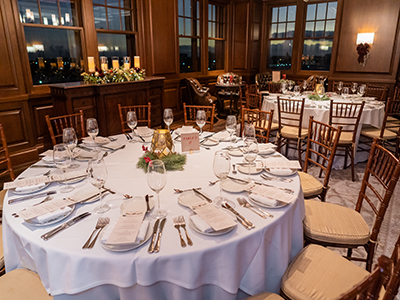 A close view of a round dining table set with folded napkins, glassware, printed menus, and a subtle holiday centerpiece near large wood-framed windows.