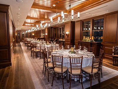A spacious banquet hall with continuous rows of round tables, wooden chairs, warm overhead lighting, and paneled walls extending throughout the room.