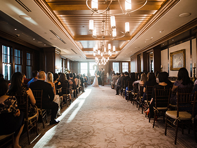 A wide view of an indoor wedding ceremony with guests seated in rows along a central aisle leading to the couple at the altar, illuminated by evenly spaced chandeliers.