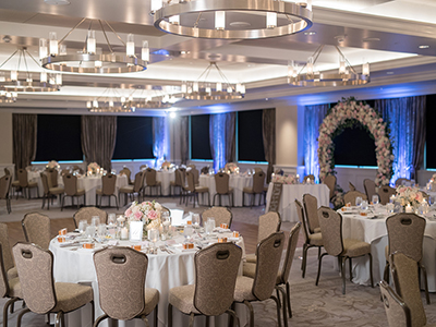 An elegant ballroom set for a wedding reception with round tables in white linens, modern ring chandeliers, soft blue uplighting, and floral centerpieces throughout the space.