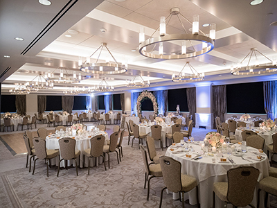 A wide view of a refined reception room featuring evenly spaced round tables, cushioned chairs, contemporary ceiling fixtures, and ambient blue accent lighting.