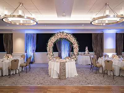 A symmetrical wedding reception setup highlighting a circular floral arch centerpiece framed by draped windows and softly lit dining tables.