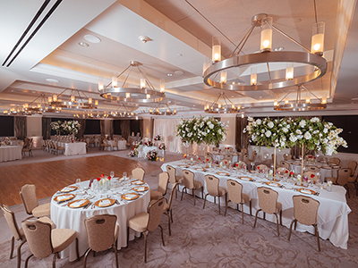 A formal reception space with long and round tables, white linens, elegant floral arrangements, warm chandelier lighting, and a polished dance floor.