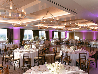 A spacious wedding reception hall featuring round tables, white linens, wooden chairs, dramatic chandelier lighting, and soft purple uplighting throughout the room.