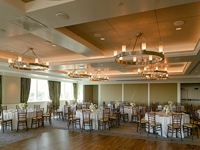 A bright banquet room arranged with round dining tables, white tablecloths, wooden chairs, and contemporary chandeliers above a wood dance floor.