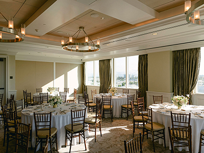 A sunlit event space with large windows, round tables set with white linens and floral centerpieces, and modern ceiling lighting creating an airy atmosphere.