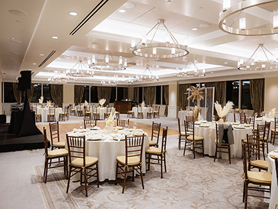 A spacious wedding reception room arranged with round tables, white linens, wooden chairs, pampas grass accents, and modern circular chandeliers overhead.