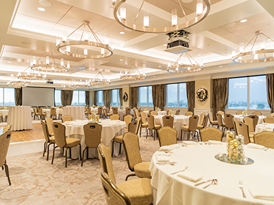 A large banquet room prepared for a formal event with round tables, white linens, modern lighting, and a presentation screen near the dance floor.