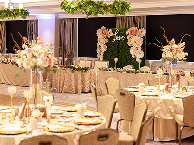 A styled sweetheart or head table set with shimmering linens, floral arrangements, greenery accents, and a decorative backdrop displaying the word “love.”