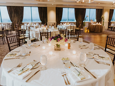 A fully set round dining table with white linens, candlelight accents, floral centerpiece, and place settings inside a bright reception hall with large windows.