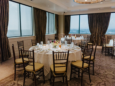 A cozy dining arrangement near large windows featuring round tables, white linens, wooden chairs, floral centerpieces, and natural daylight views.