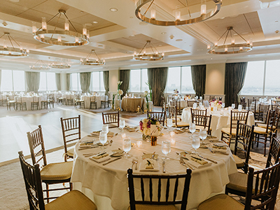 A wide view of a light-filled reception space with round dining tables, white linens, wooden chairs, and modern chandeliers above a wood dance floor.