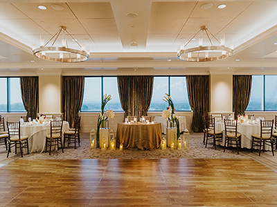 A symmetrical reception setup showcasing a central sweetheart table flanked by lanterns, candles, and floral accents against tall draped windows.