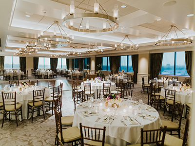 A panoramic view of an elegant ballroom with evenly spaced round tables, white linens, wooden chairs, and contemporary chandelier lighting.