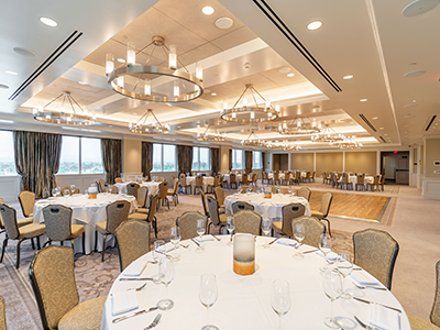 A bright and elegant banquet room with round tables set for dining, gold chairs, modern circular chandeliers, and large windows along the walls.