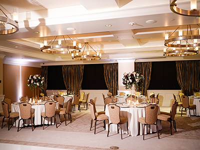An elegant banquet space featuring round tables with white tablecloths, tall floral centerpieces, cushioned chairs, and warm chandelier lighting throughout the room.