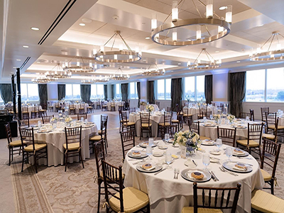 A formal reception setup with round tables dressed in white linens, place settings, floral centerpieces, and dark wood chairs beneath modern chandeliers.