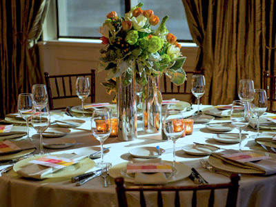 A round table set with glassware and flatware, green, white, and orange flowers and candles.