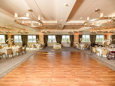 A softly lit evening reception space featuring round tables, white linens, simple floral centerpieces, wooden chairs, and large windows framing an outdoor view.