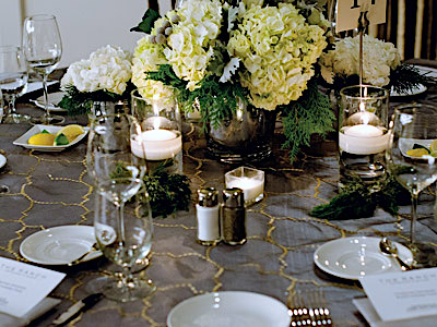 A round table set with glassware and flatware, green and white flowers, and candles.