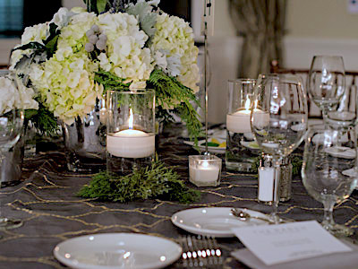 A closeup of a round table set with glassware and flatware, green and white flowers, and candles.