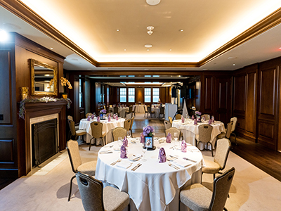 A formal dining room with round tables, white tablecloths, a fireplace feature, soft overhead lighting, and elegant table settings for an event.