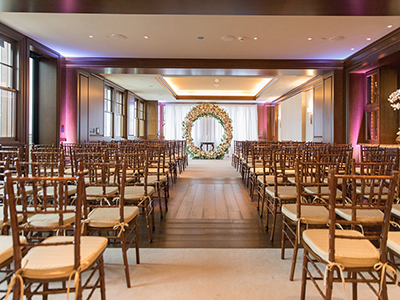 An empty indoor ceremony space prepared with rows of wooden chairs, a central aisle, and a large floral wreath positioned at the front beneath warm ambient lighting.