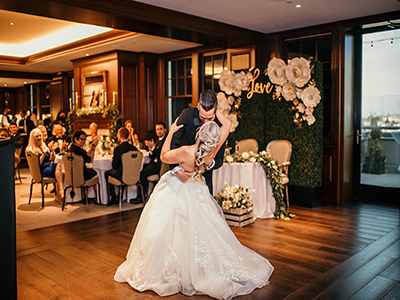 A couple sharing a first dance on a wood floor in an elegant reception room with round dining tables, floral décor, and a greenery backdrop reading “love.”