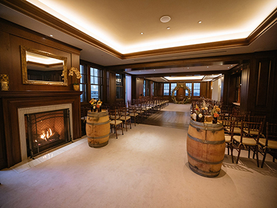 A wide view of an indoor ceremony room featuring a fireplace, barrel décor at the entrance, rows of wooden chairs, and a floral arch at the far end of the aisle.