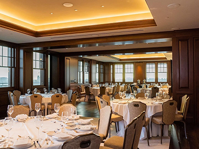 A refined event space featuring round tables set with glassware and linens, wood floors, large windows, and warm ambient ceiling lighting.