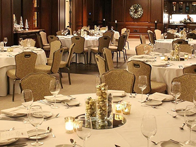 A holiday-decorated banquet room with round tables, candlelit centerpieces, wood-paneled walls, and a softly lit bar area in the background.