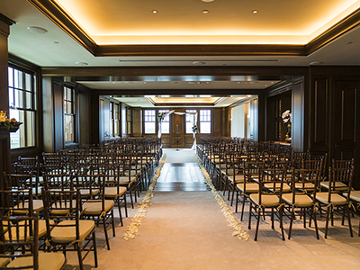 A formal indoor ceremony space with a long aisle lined with white petals, symmetrical rows of wooden chairs, and soft uplighting highlighting the ceiling and walls.
