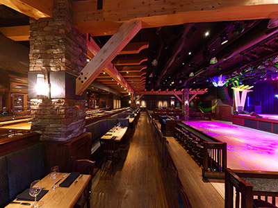 A view from the right side of the Ranch Saloon showing a wooden dance floor, stage, guitar disco ball, tables and chairs, and a bar.