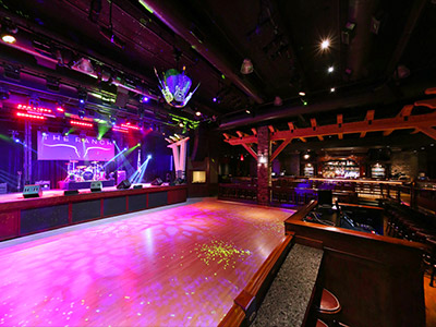 A view from the left side of the Ranch Saloon showing a wooden dance floor, stage, guitar disco ball, tables and chairs, and a bar.