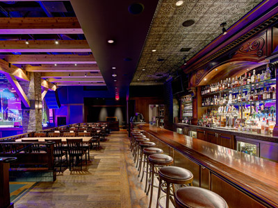 A bar with stools, next to wooden tables and chairs overlooking a dance floor and stage