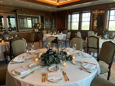 A round dining table elegantly set with white linens, gold flatware, folded napkins, glassware, candlelight, and a lantern centerpiece surrounded by greenery in a wood-paneled private dining room.