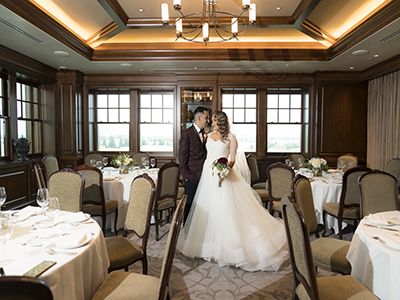 A wedding couple standing together in an intimate reception room filled with round dining tables, white linens, upholstered chairs, and large windows framed by rich wood paneling.


