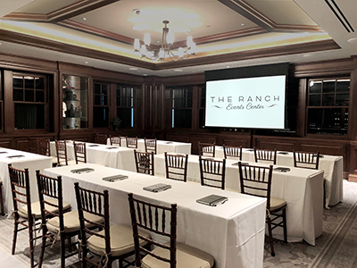 A classroom-style meeting setup with long tables, white linens, chairs arranged in rows, notepads at each seat, and a projection screen at the front of the room.