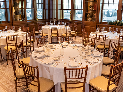 A bright and polished dining room arranged for a formal event, featuring round tables, white linens, wooden chairs, and large windows filling the space with natural light.